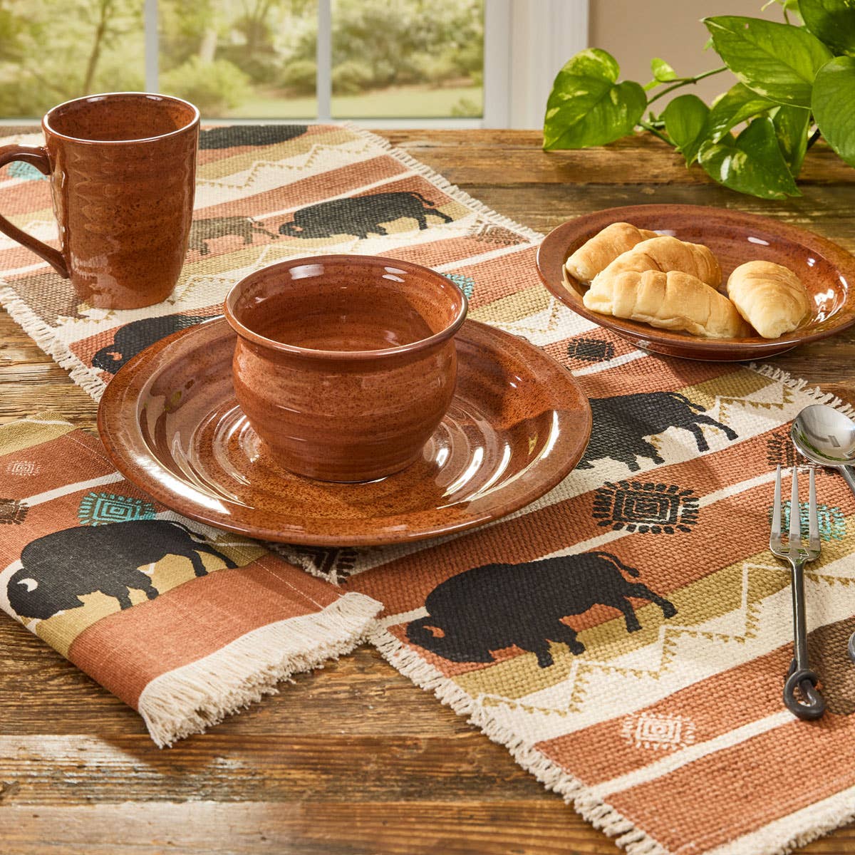 "Park Designs Sinclair Tomato Soup cereal bowl and salad plate styled on a rustic wood table with a Southwest-style bison table runner, matching mug, croissants, and silver cutlery"