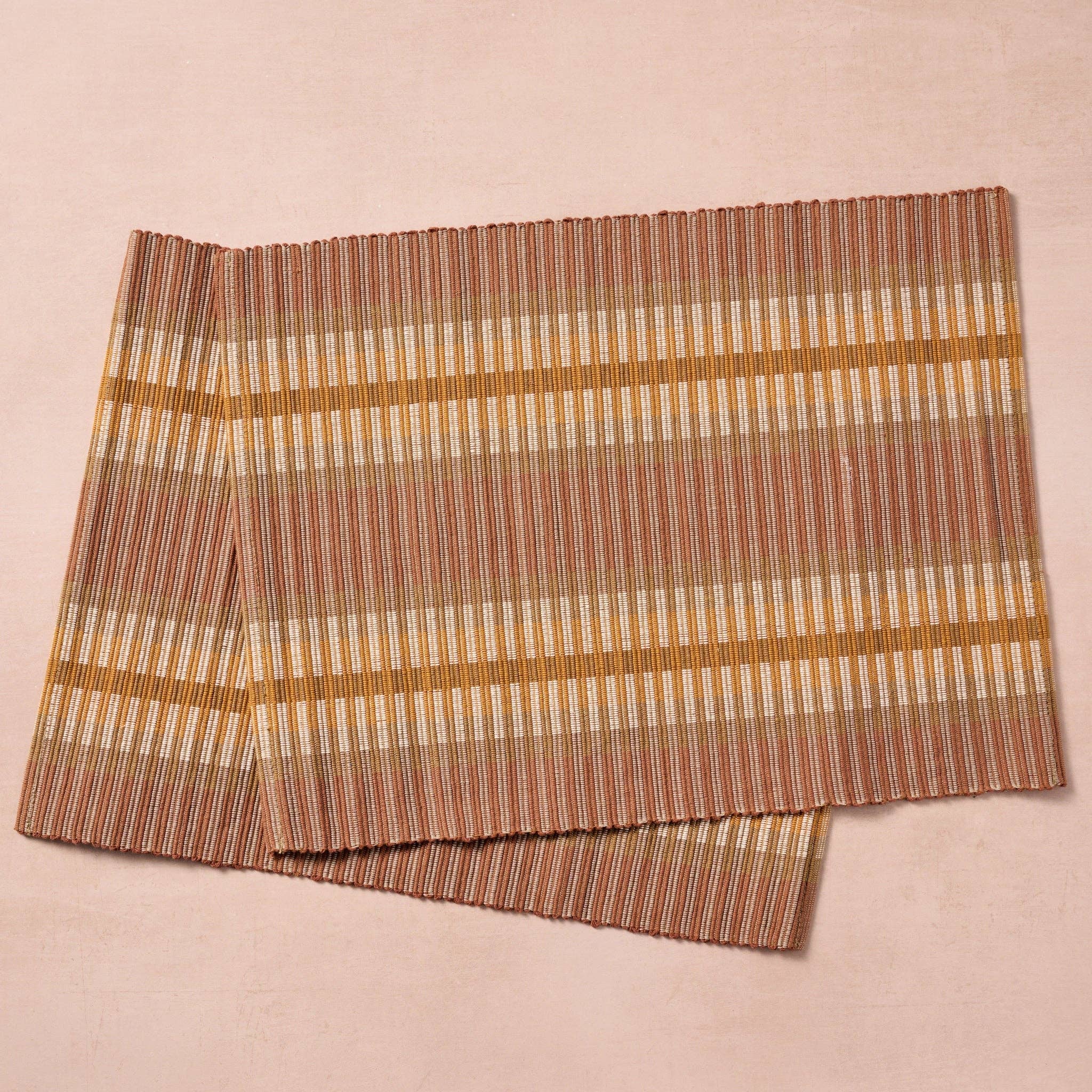 Two placemats fanned/overlapping (blush bg): "Two handloomed 100% cotton placemats in brown, tan, and gold stripe, handcrafted in Nepal by Ten Thousand Villages, shown overlapping on a blush background"