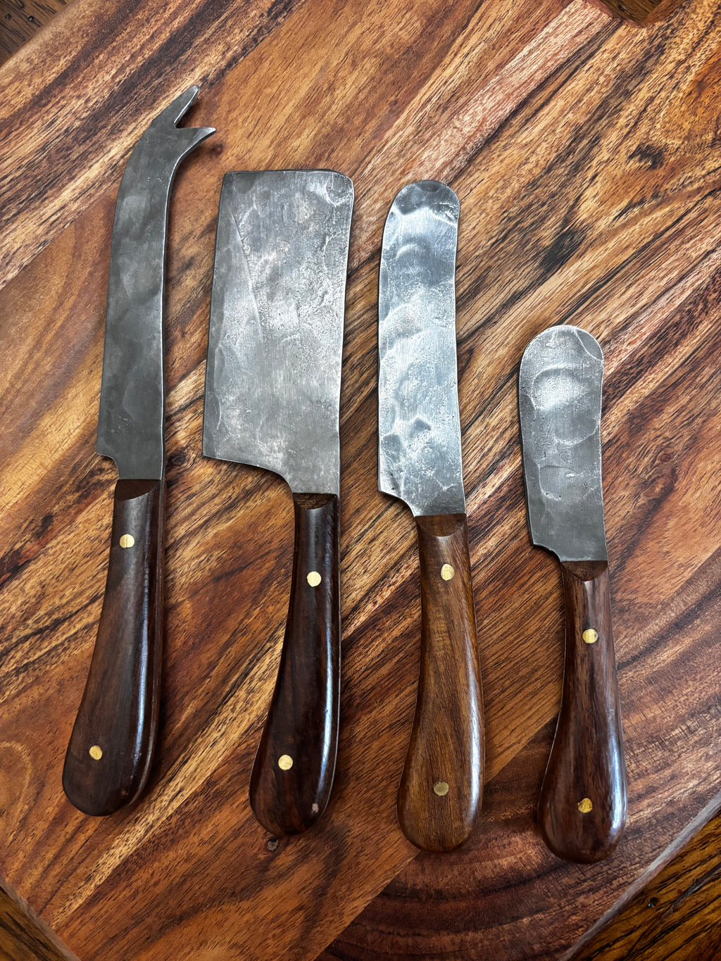 All 4 knives laid out on walnut board, overhead → "Set of four Farmhouse Pottery artisan forged charcuterie cheese knives with hammered steel blades and dark walnut handles with brass rivets, laid out on a walnut wood board overhead view"