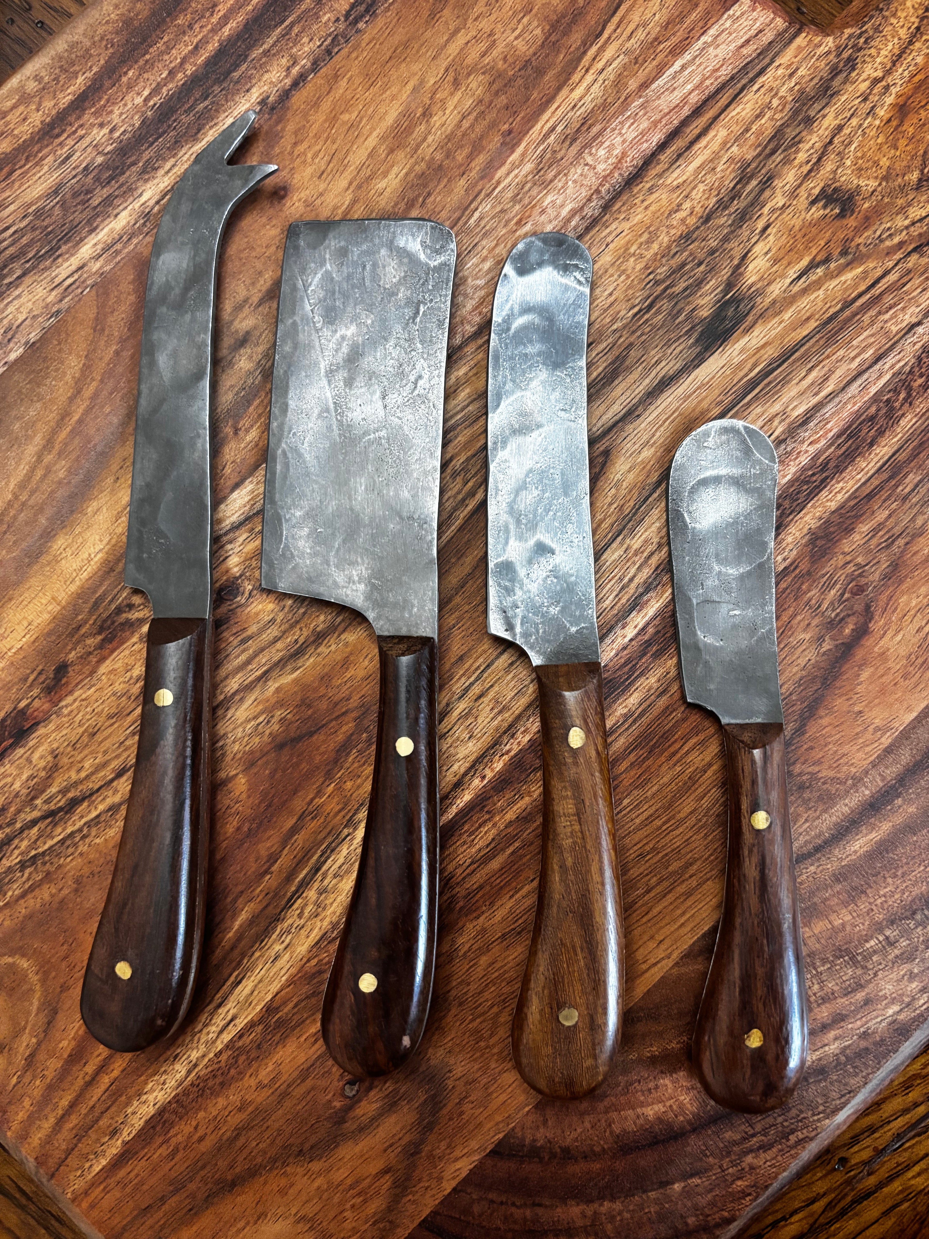 All 4 knives laid out on walnut board, overhead → "Set of four Farmhouse Pottery artisan forged charcuterie cheese knives with hammered steel blades and dark walnut handles with brass rivets, laid out on a walnut wood board overhead view"