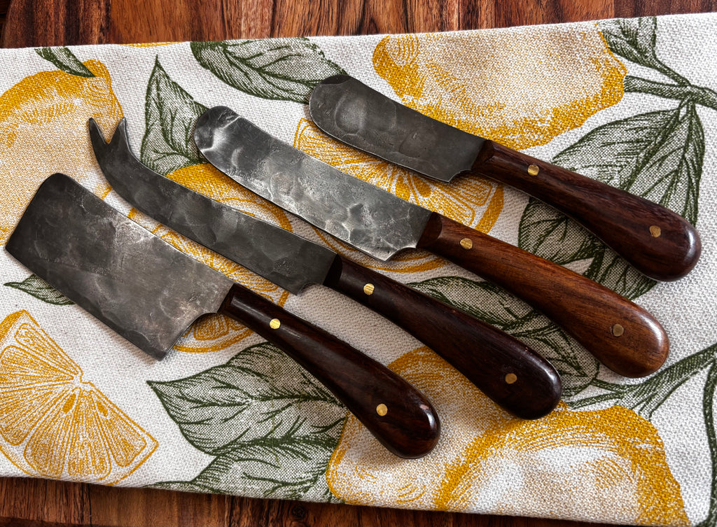 All 4 knives on lemon print linen, overhead → "Set of four Farmhouse Pottery artisan forged cheese knives with hammered steel blades and walnut handles displayed on a yellow lemon print linen kitchen towel on a dark wood surface"