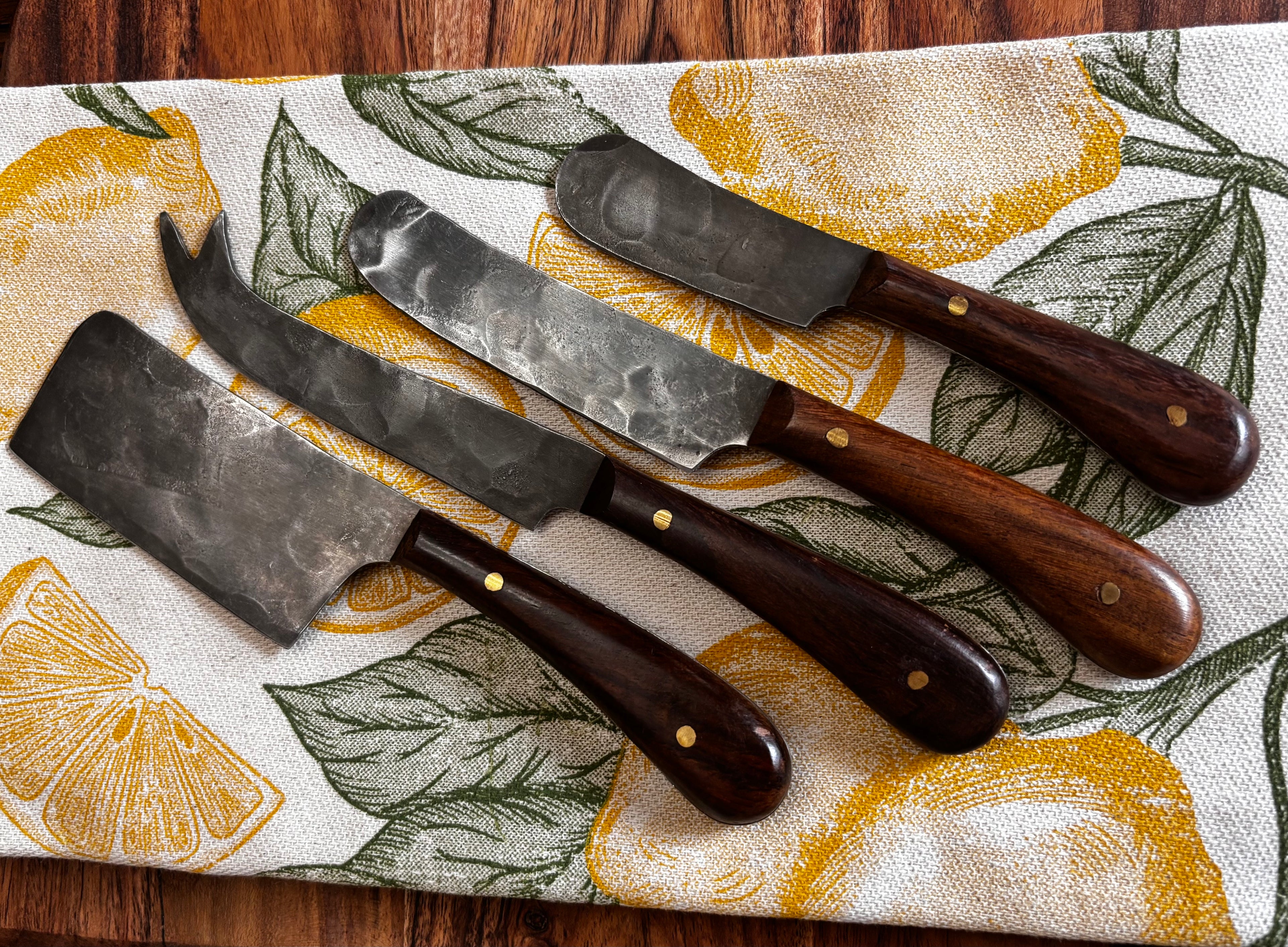 All 4 knives on lemon print linen, overhead → "Set of four Farmhouse Pottery artisan forged cheese knives with hammered steel blades and walnut handles displayed on a yellow lemon print linen kitchen towel on a dark wood surface"
