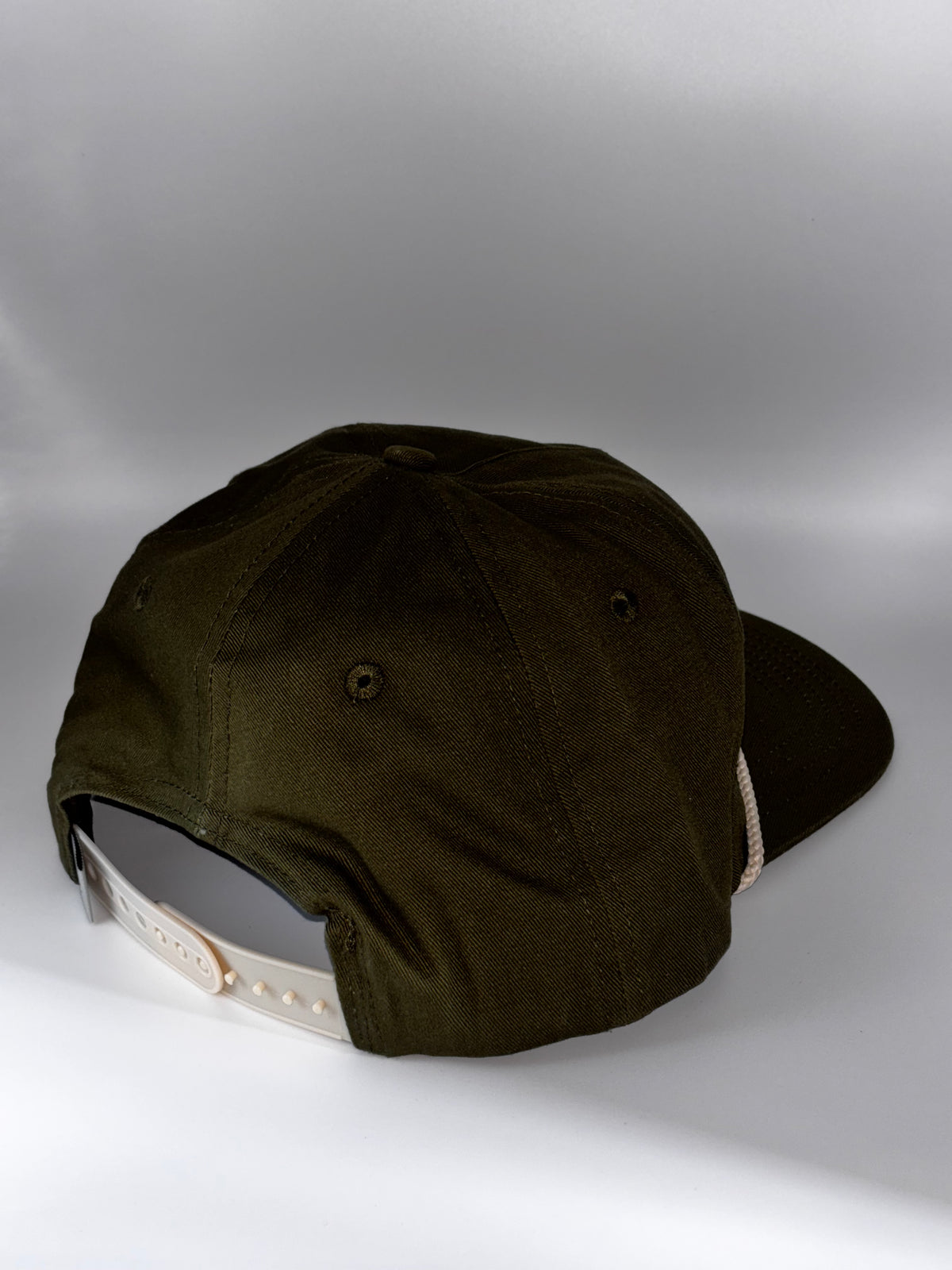 An olive green baseball cap shown from the back, featuring a cream-colored adjustable strap and a dark visor, set against a neutral gray studio background.