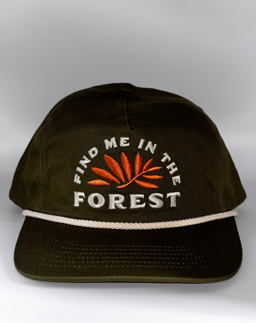 An olive green baseball cap with white embroidered text reading 'FIND ME IN THE FOREST' and an orange leaf design on the front, plus a white rope detail across the brim, shown against a neutral gray background.