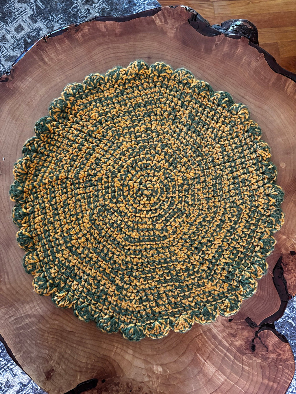 A round, hand-crocheted cat lounger with a scalloped edge, featuring a variegated pattern of dark green and mustard yellow. The placemat is displayed on a rustic live-edge wood slice, with a patterned textile and a wooden floor visible in the background.