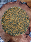 A round, hand-crocheted cat lounger with a scalloped edge, featuring a variegated pattern of dark green and mustard yellow. The placemat is displayed on a rustic live-edge wood slice, with a patterned textile and a wooden floor visible in the background.