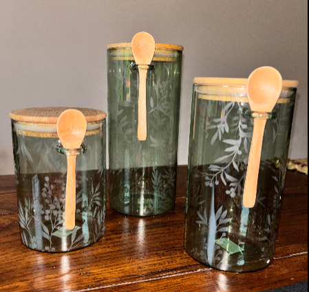 Set of three sage green floral etched glass canisters in graduated sizes with natural bamboo lids and wooden spoons, displayed on dark wood surface — The Everyday Shoppe"