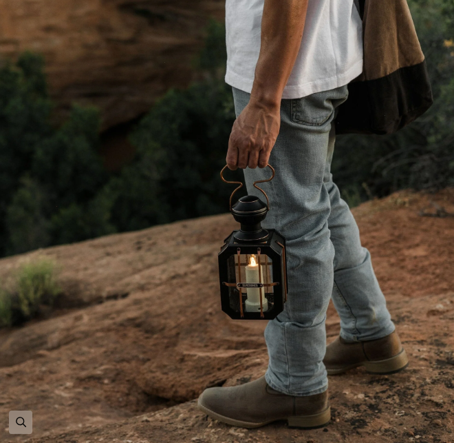 Person carrying Barebones Box Candle Lantern with lit candle while walking on rocky terrain outdoors with canyon landscape in background