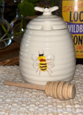 Lifestyle — honey pot with dipper, wildberry honey jar, plants in background: "White porcelain beehive honey pot with bee detail and wooden honey dipper on a woven surface beside a jar of local wildberry honey with green plants in the background"
