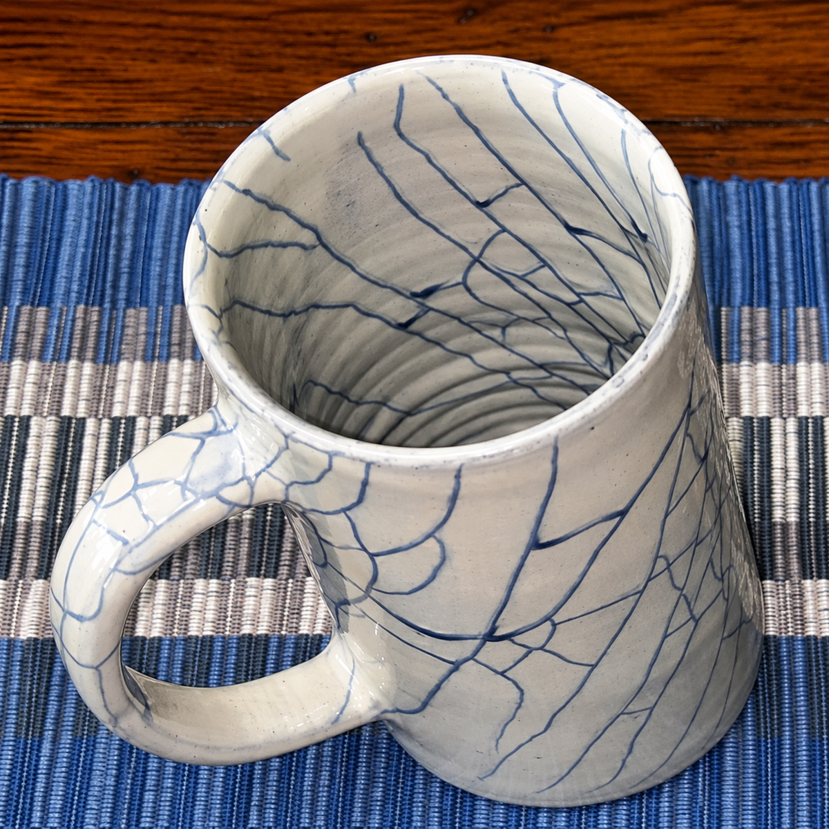 Overhead view of Indigo Night Blue Crackle handcrafted stoneware mug showing intricate hand-painted crackle glaze detail inside and out, resting on blue striped handloomed placemat — Ten Thousand Villages fair trade — Ramen Night In Collection"