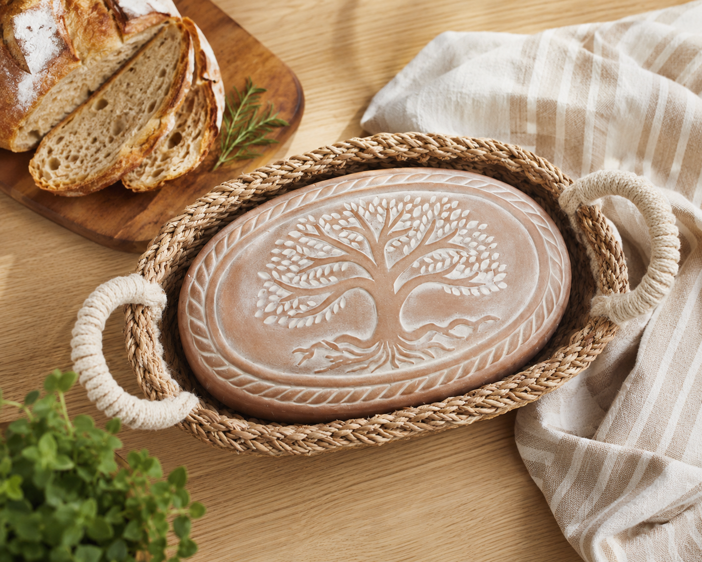 "Korissa oval terracotta bread warmer with carved Tree of Life design nestled in a seagrass basket with rope handles on a wood table beside sliced artisan sourdough on a cutting board, fresh herbs, and a striped linen towel"