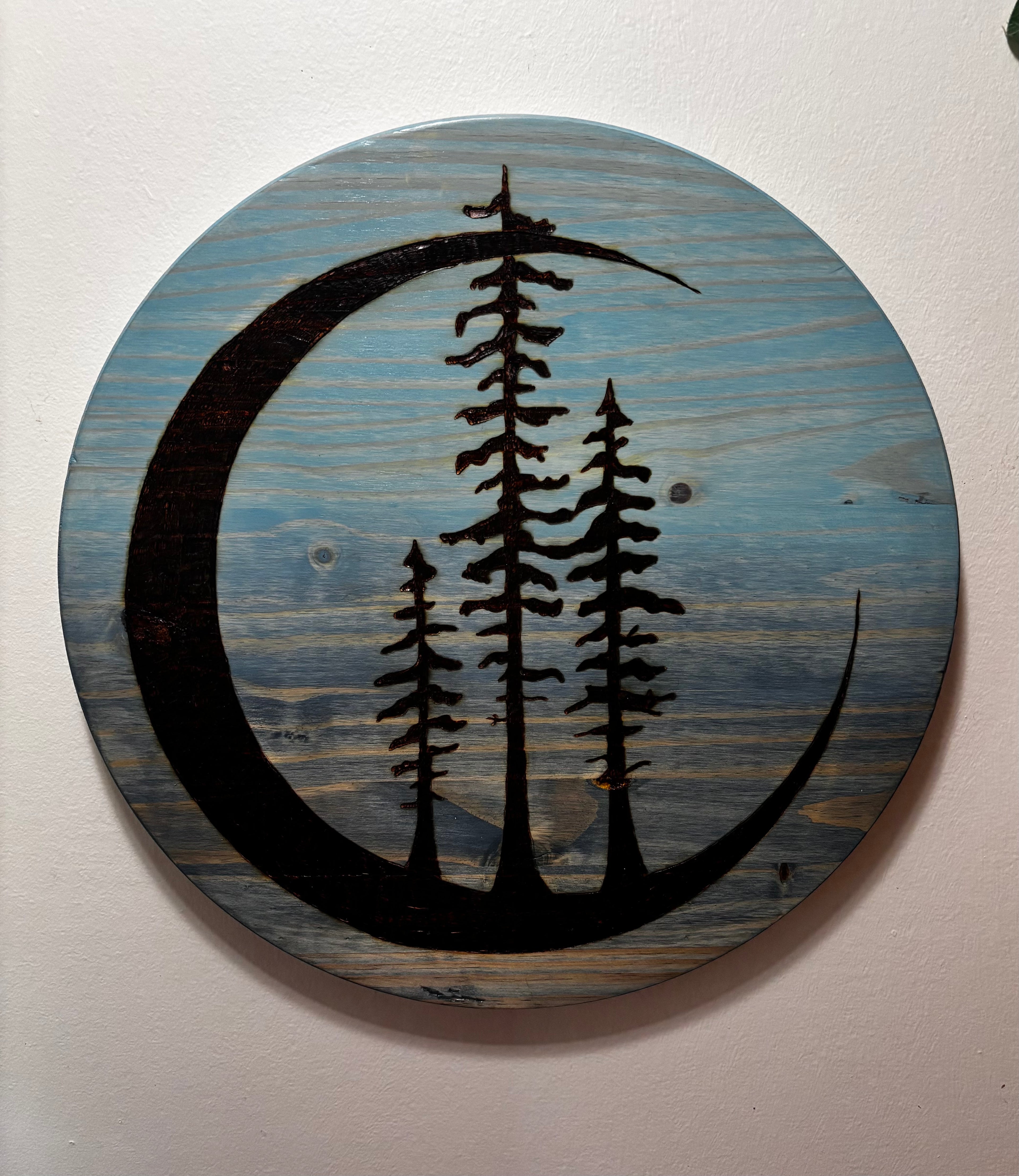  clean white wall, full front view): "Tres Luna crescent moon and pine tree pyrography and painted wall art on round pine, 18 inches, displayed on white wall — Adirondack Art Co. by Darren Segool"