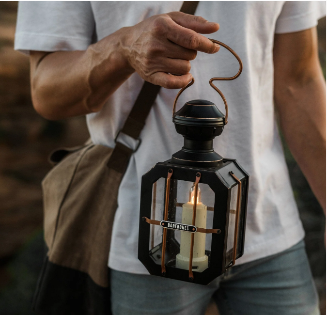 Person carrying Barebones Box Candle Lantern by copper handle with lit candle inside, wearing white t-shirt and canvas bag outdoors"