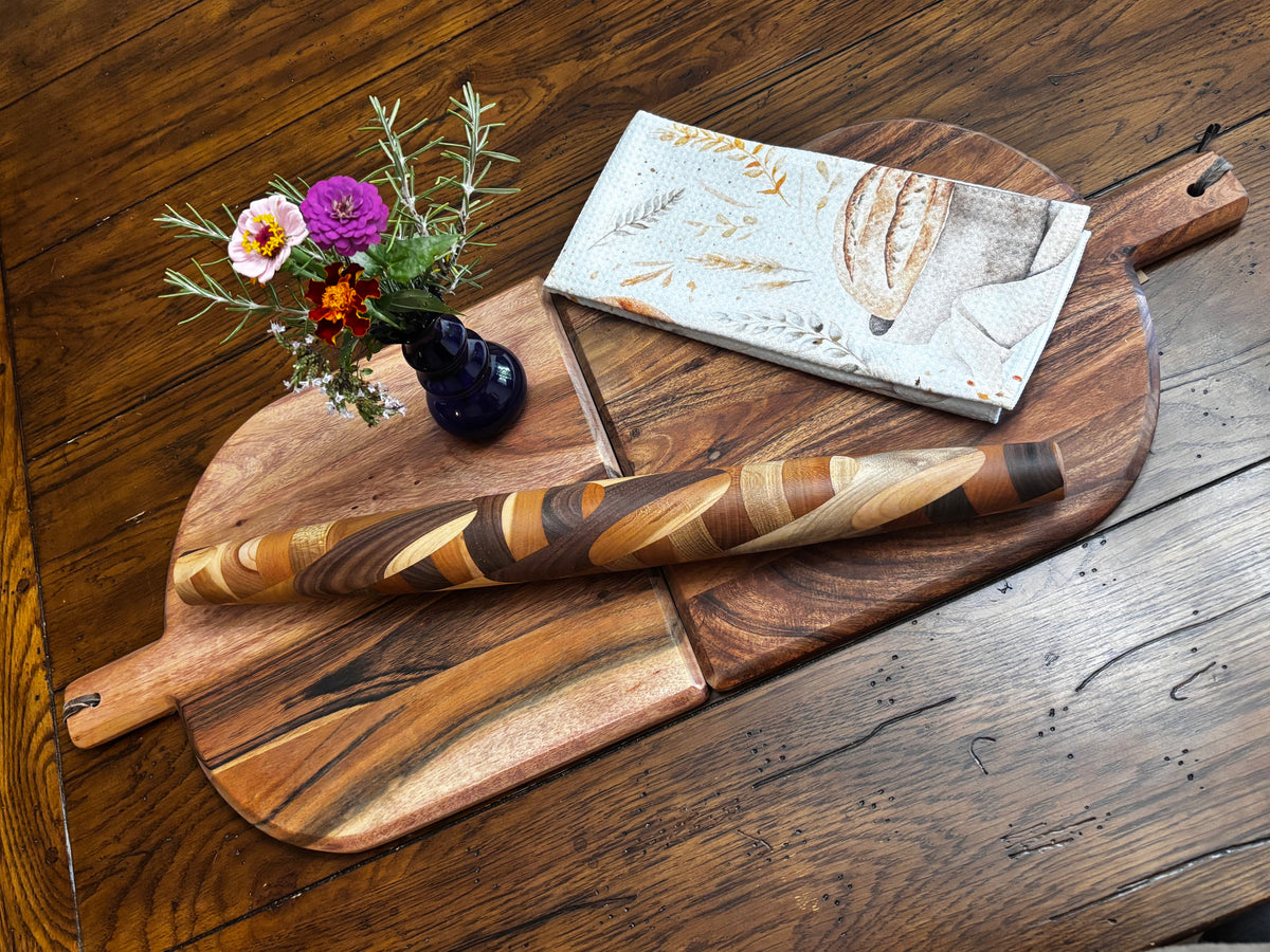 Wide lifestyle shot, full rolling pin across two boards (round + paddle style) with blue bud vase and bread towel, rustic wood table. "Miss Clarissa French rolling pin, mixed Black Walnut, Cherry and Maple hardwood, styled on artisan wood boards with fresh flowers — handmade in Tennessee"