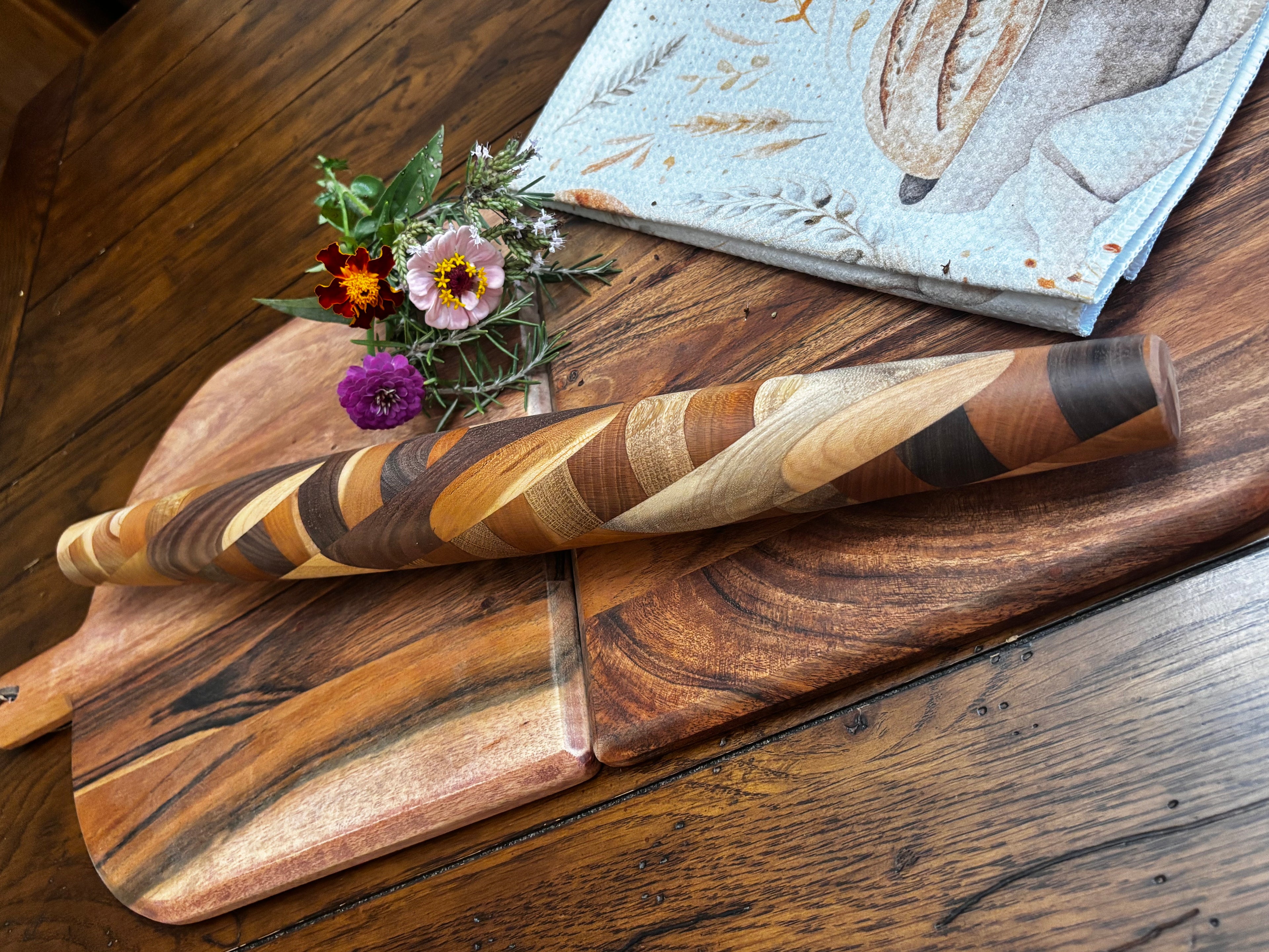Close-up angled shot showing the beautiful spiral wood grain detail of the rolling pin across boards with fresh flowers. "Close-up detail of handmade French rolling pin showing swirling mixed hardwood grain — Black Walnut, Cherry and Maple — The Rileyland Collection"