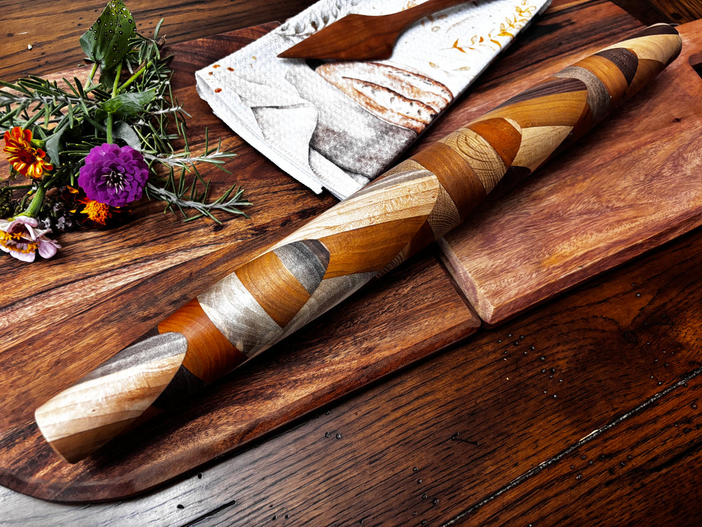  Dramatic close-up of the tapered end showing the intricate wood inlay pattern, with flowers and bread towel in background. "Detail shot of tapered end of handmade mixed hardwood French rolling pin showing intricate spiral wood inlay pattern — handmade in rural Tennessee"
