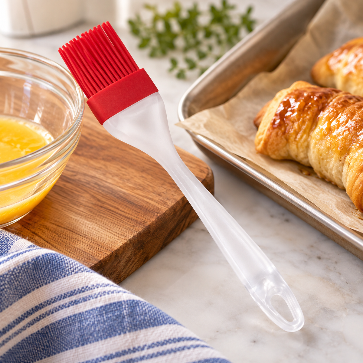 Mrs. Anderson's 8-inch red silicone pastry brush resting on a wood cutting board beside a glass bowl of egg wash and a baking sheet of freshly glazed croissants on parchment paper.