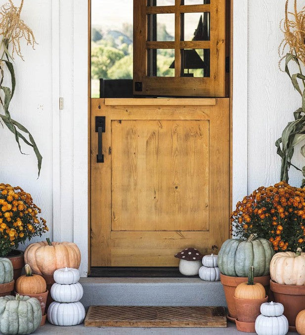 Lifestyle — fall porch with pumpkins and mums → "Wool felted mushroom doorstop with sage green polka dot cap displayed on a farmhouse porch step surrounded by fall pumpkins, orange mums, and dried corn stalks beside a wooden Dutch door"