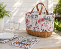 A floral picnic basket with a wicker base and brown leather handles sits on a wooden table outdoors, styled with plates, wine glasses, cutlery, and a potted plant.