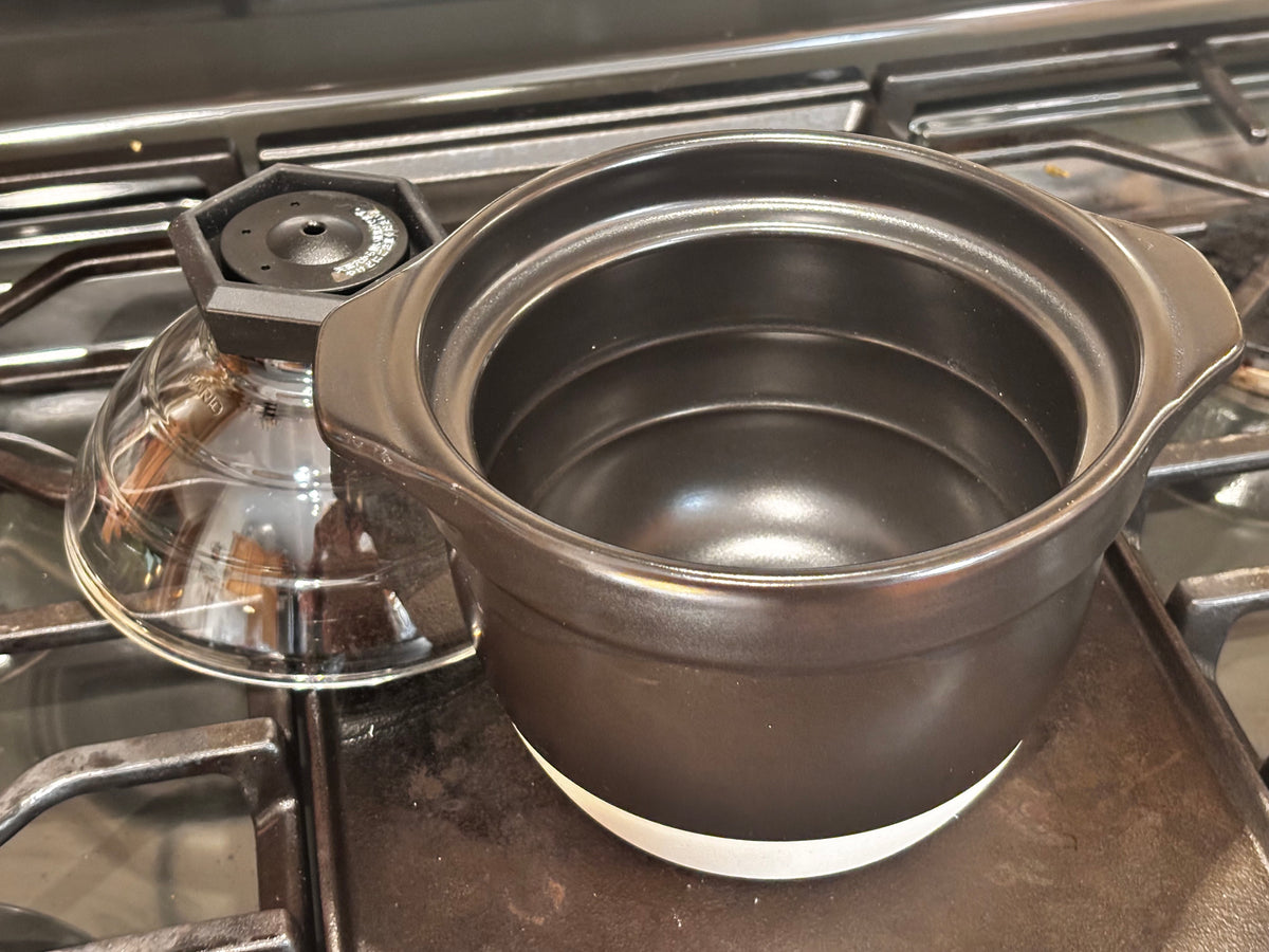 Hario Gohan Gama ceramic rice cooker with lid removed showing empty black ceramic interior, sitting on gas stovetop — glass dome lid with faceted knob resting beside it"