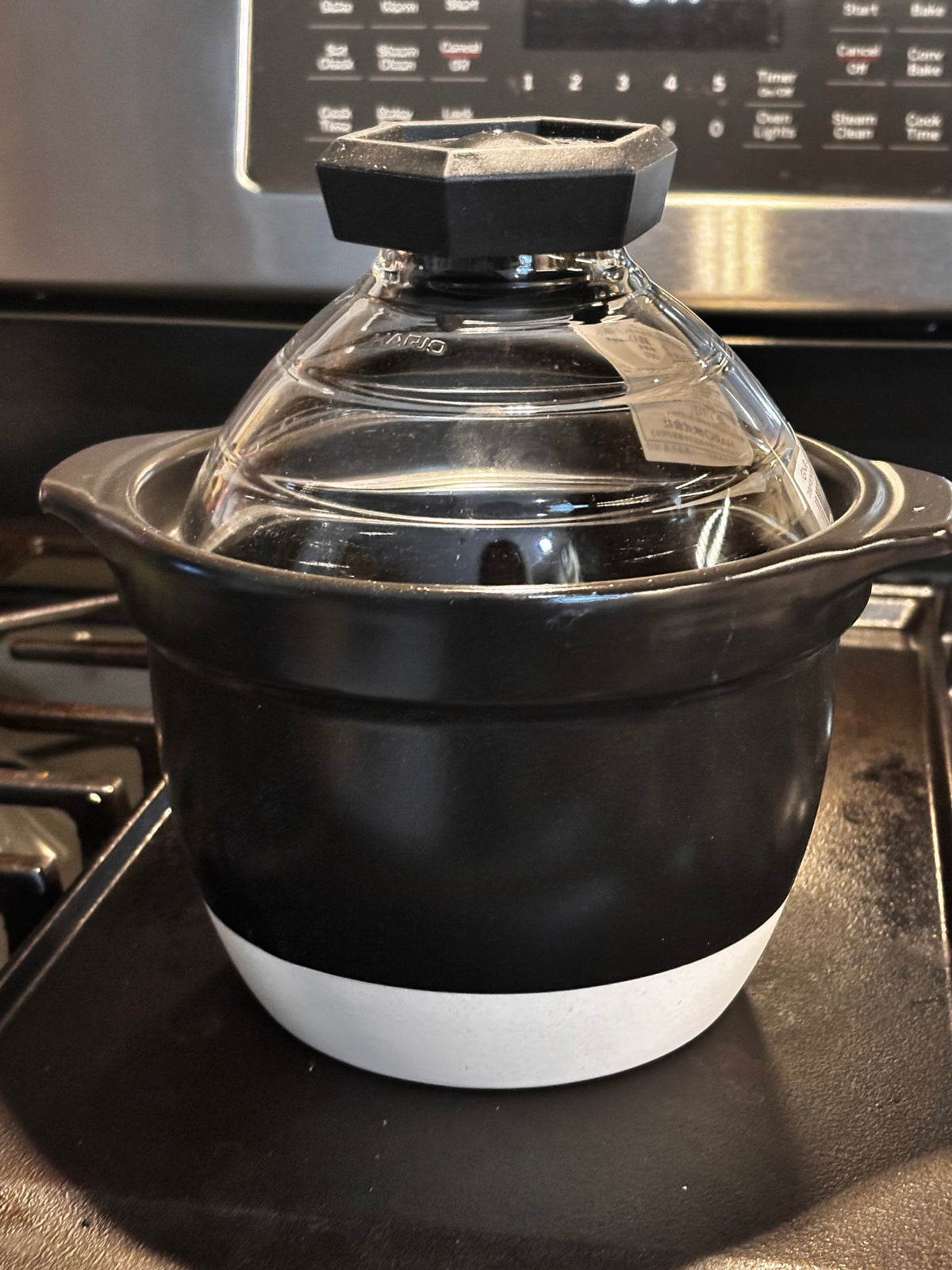 rice_cooker_2 — lid on, on stovetop with oven in background): "Hario Gohan Gama ceramic rice cooker with glass dome lid on gas stovetop, ready to cook — black ceramic body with white base band and faceted glass lid knob, stainless oven in background"