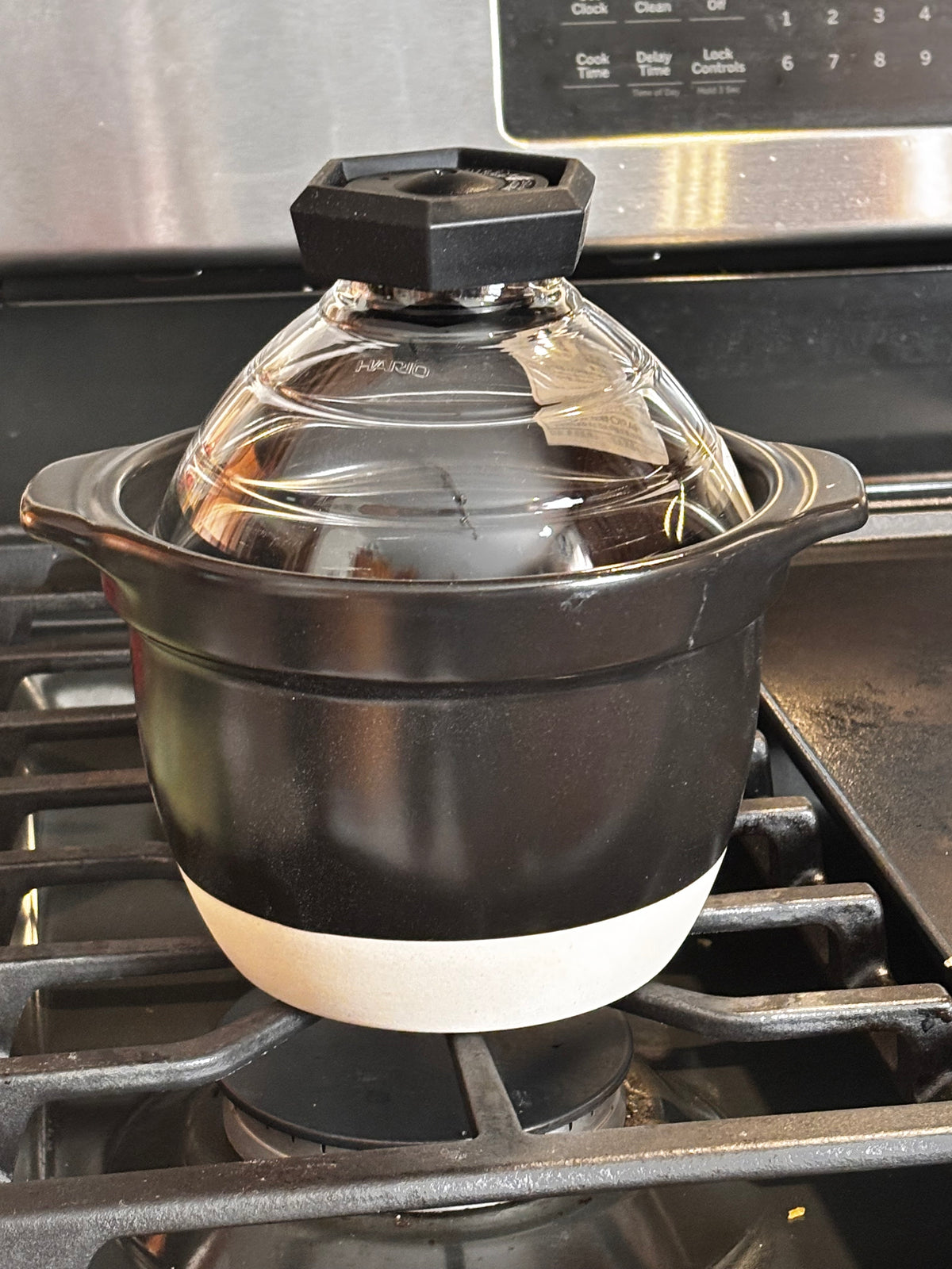2 — lid on, on stovetop with oven in background): "Hario Gohan Gama ceramic rice cooker with glass dome lid on gas stovetop, ready to cook — black ceramic body with white base band and faceted glass lid knob, stainless oven in background"
