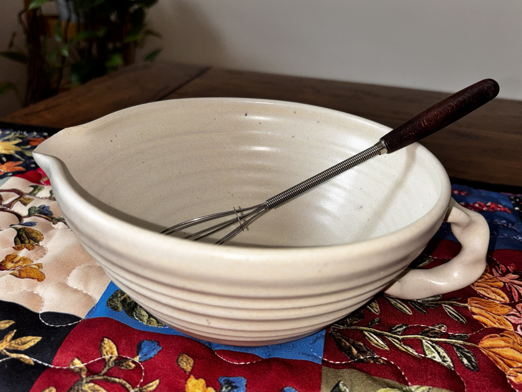  "Straight-on shot of two Gravesco handmade cream ceramic mixing bowls with dark-handled whisks on a colorful quilted table runner"