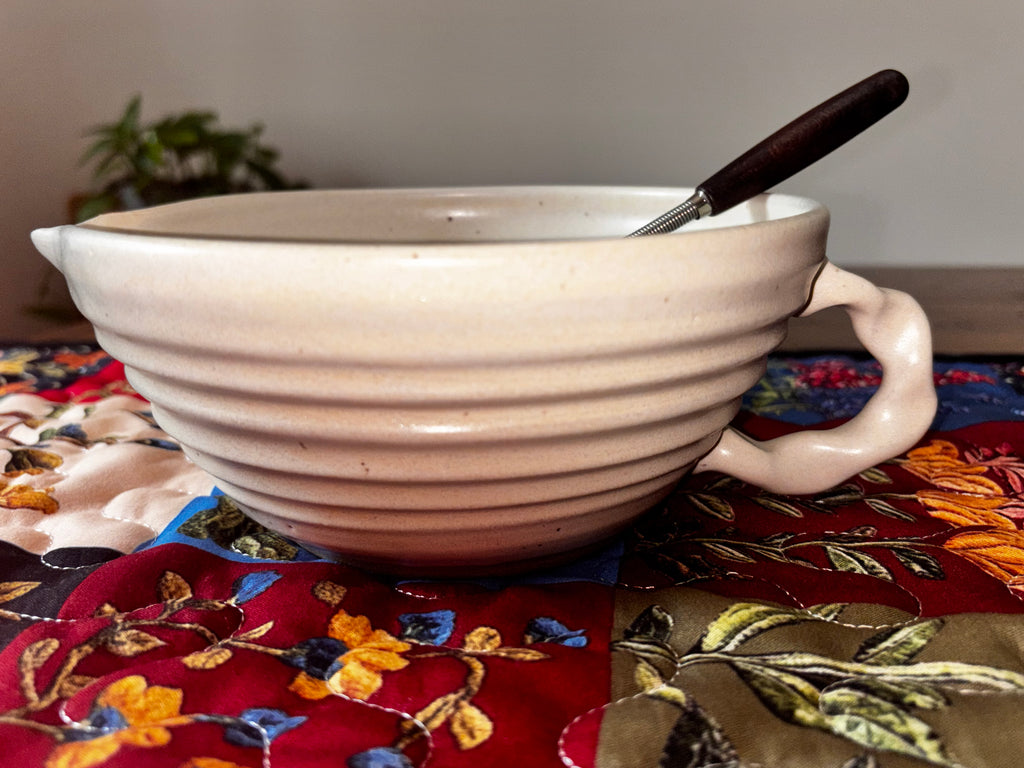 Single Gravesco handmade cream ceramic mixing bowl with pour spout, handle, and whisk on colorful quilted runner, side profile"
