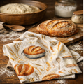(floured wood table with sourdough boule): Sourdough waffle weave kitchen towel with watercolor bread and wheat design folded on a floured rustic wood table beside a freshly baked sourdough boule on a cutting board and a wooden proofing bowl with dough