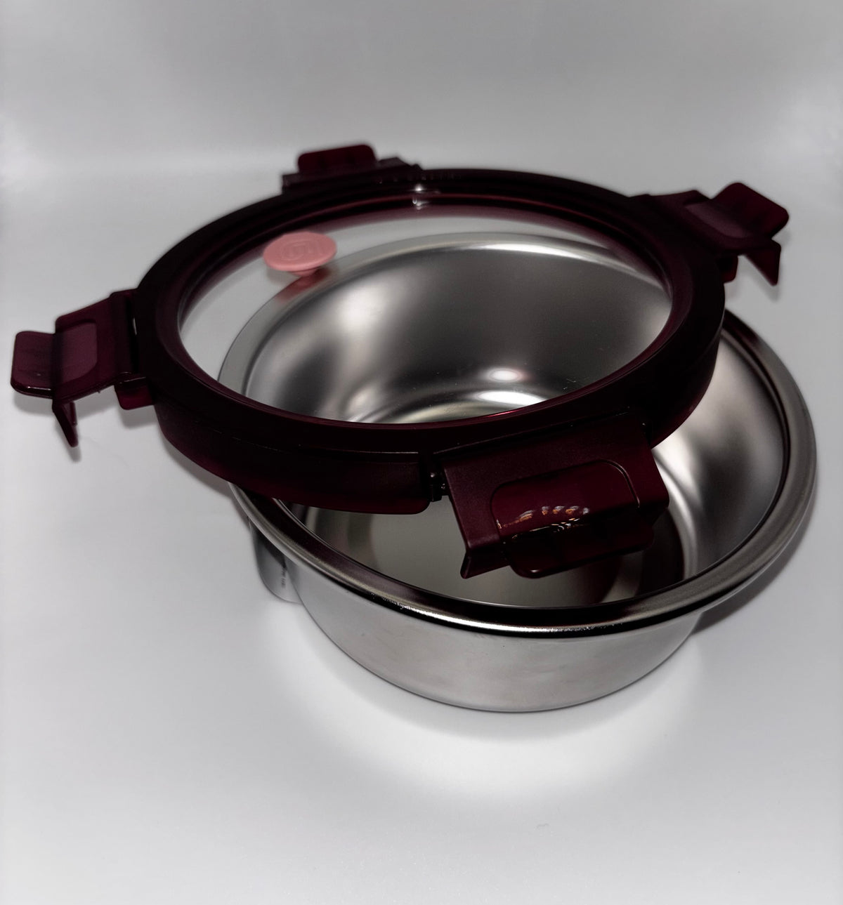 Genicook stainless steel container with its glass lid resting at an angle and burgundy locking clips open.