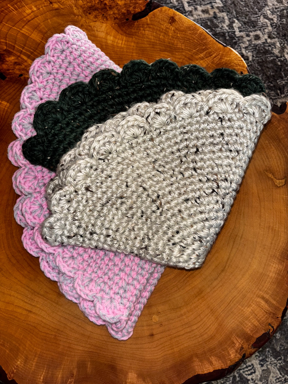 Three handcrafted crochet cat loungers in pink and gray, deep green, and natural tweed folded and stacked on a live-edge wood slab, showing the scalloped edge detail by Sleeping Whiskers Studio.