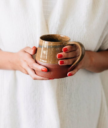 Lifestyle — woman in white holding mug with red nails: "Woman in white linen holding a handmade terracotta ceramic mug with both hands, showcasing the earthy glaze and natural terracotta base"
