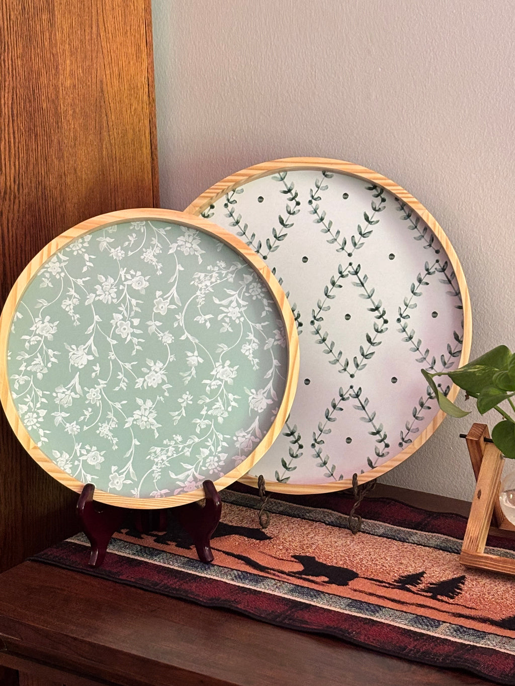 "Two round botanical print wooden trays displayed on dark wood sideboard — smaller sage green floral tray in front, larger white diamond lattice leaf print tray behind — with cream green floral pitcher vase — The Everyday Shoppe"