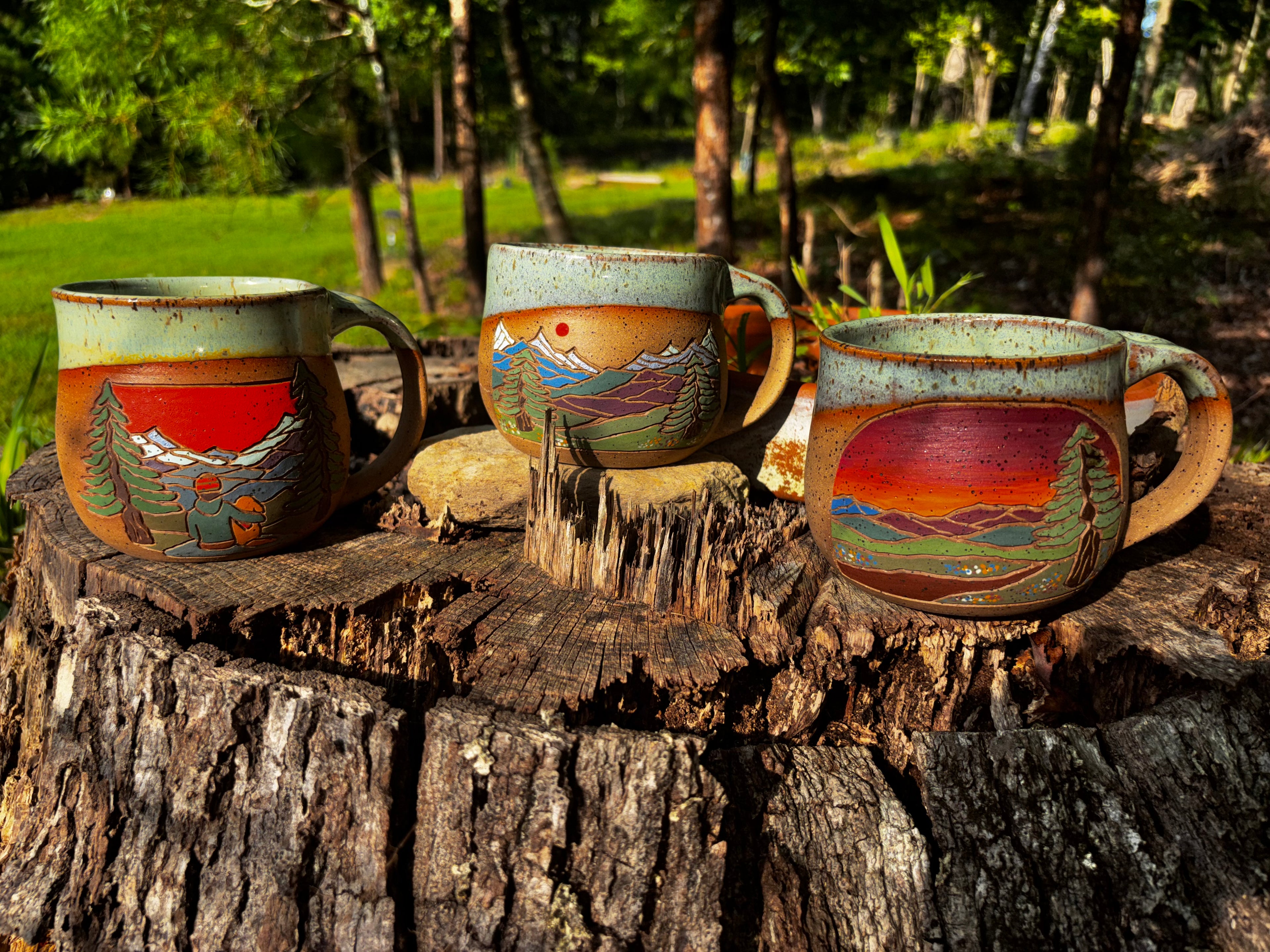 Three Forest Jeannie Pottery handmade stoneware mountain mugs displayed on a weathered tree stump outdoors, showing Trailside Friends, Quiet Ridge, and Evening Trail hand-carved landscape designs in a sunlit forest setting