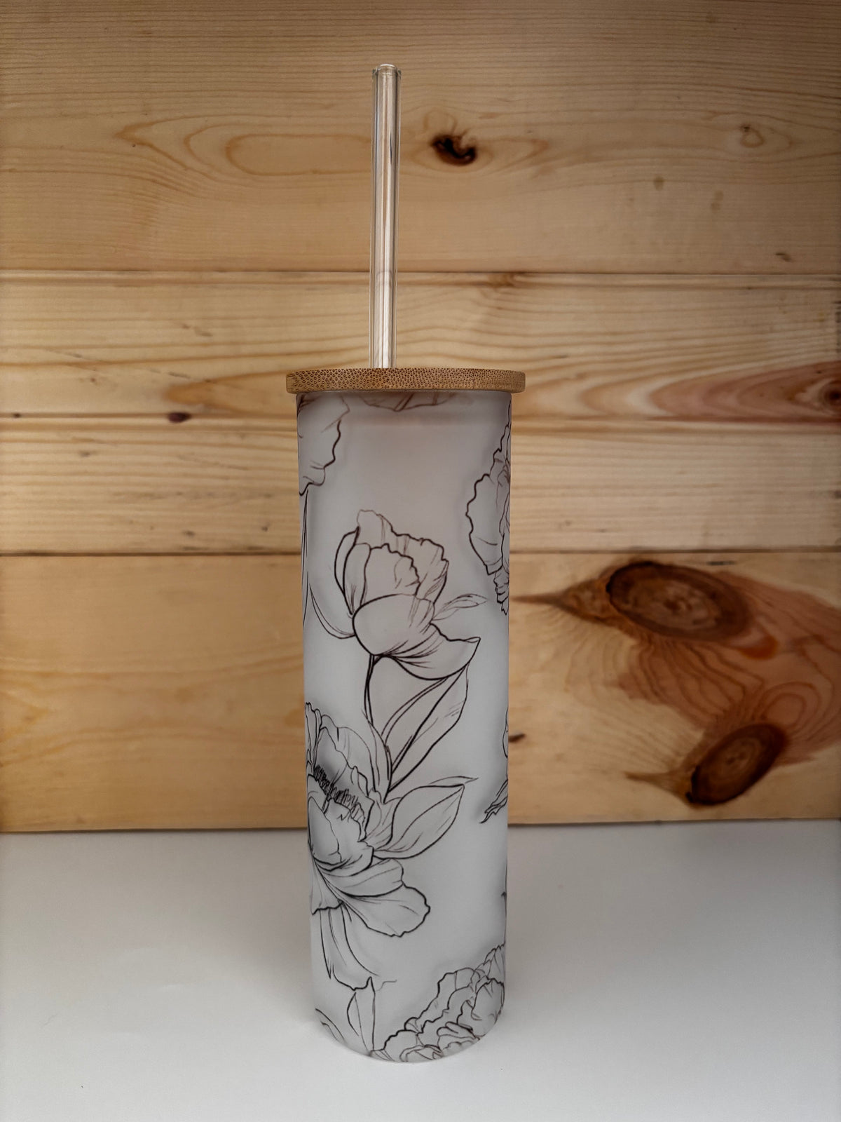 Frosted floral glass tumbler with bamboo lid and clear glass straw, displayed against a natural wood backdrop.