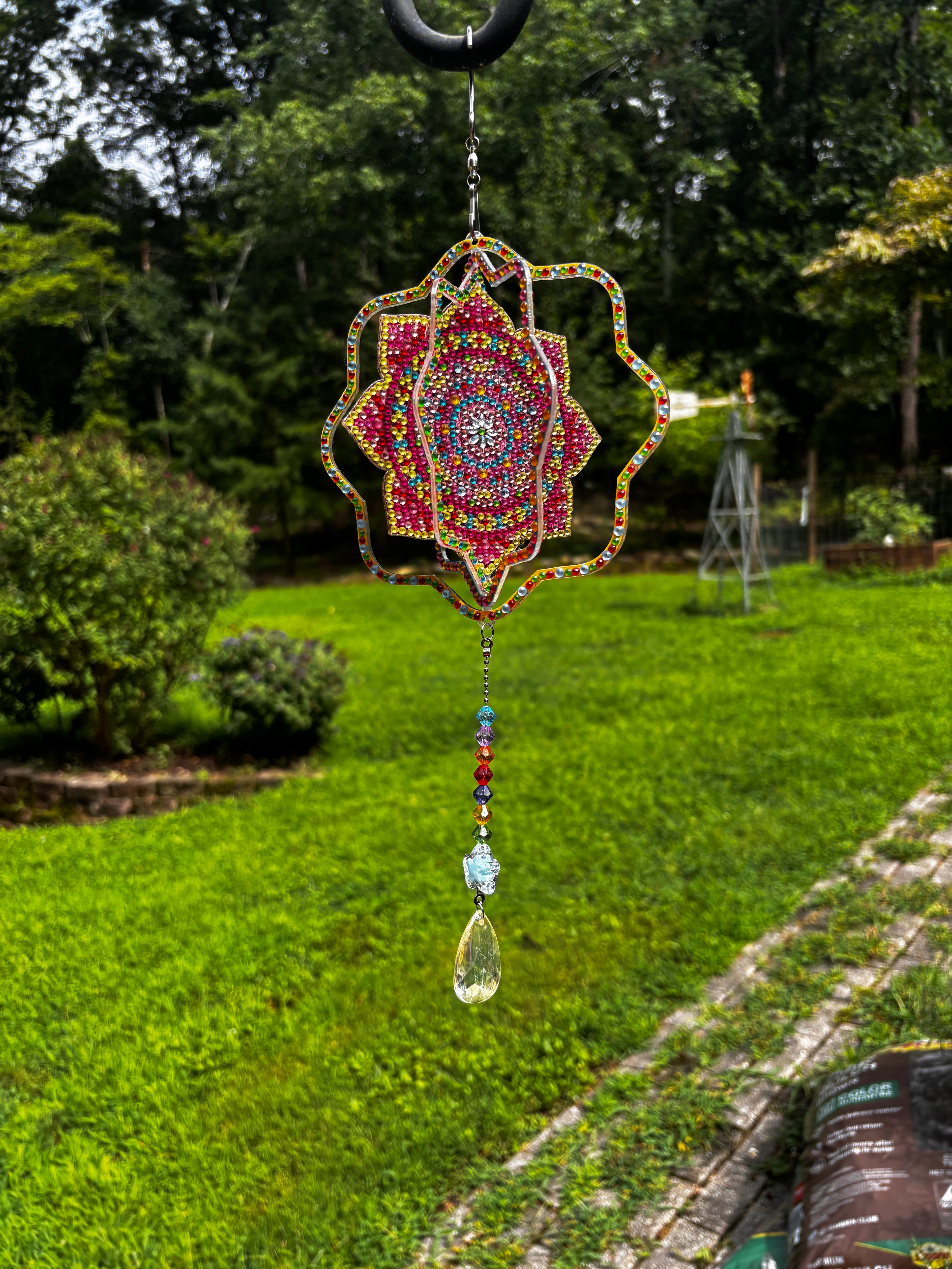 Handcrafted mandala wind spinner hanging in a backyard garden, showing the layered acrylic mandala design with colorful faceted bead strand and prism drop — by Sleeping Whiskers Studio